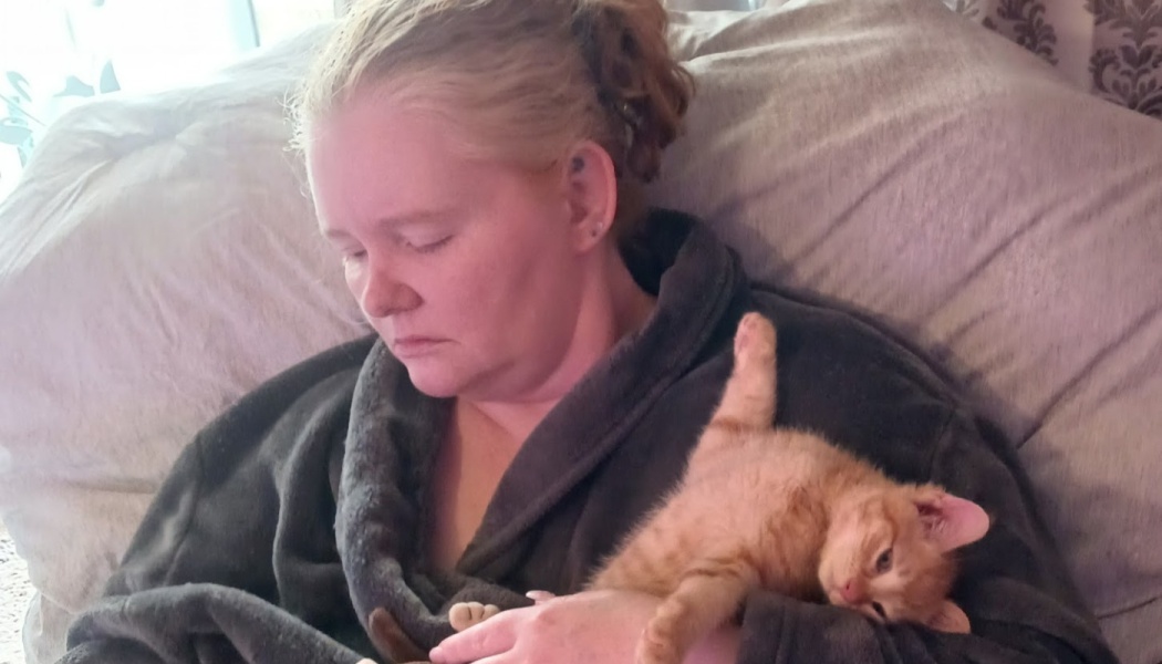 A woman in a dark fleece robe cuddles a playful orange tabby kitten stretching in her arm.