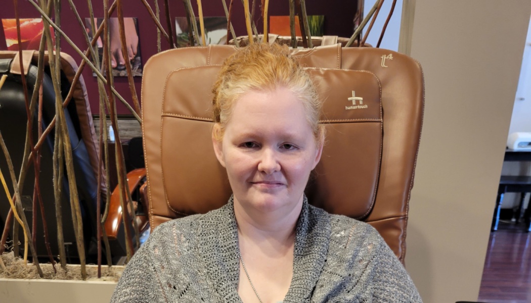 A woman with strawberry blonde hair wearing a grey sweater, smiling gently while sitting in a massage chair.