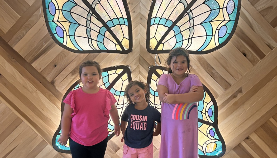 Three young girls smiling in front of large, colorful stained-glass butterfly wings mounted on a wooden wall.