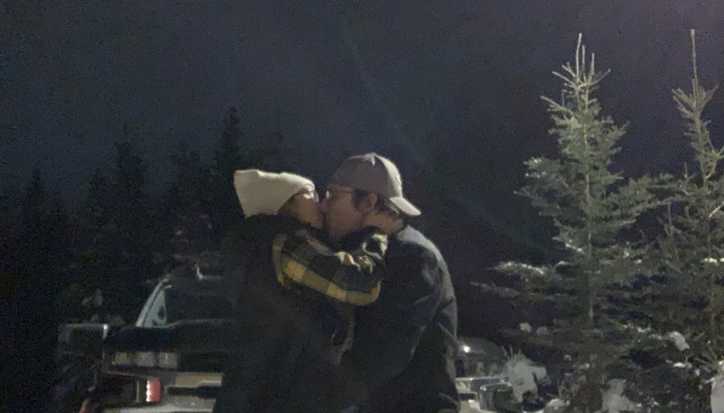 A couple wearing winter hats kissing outdoors at night near pine trees and a truck.
