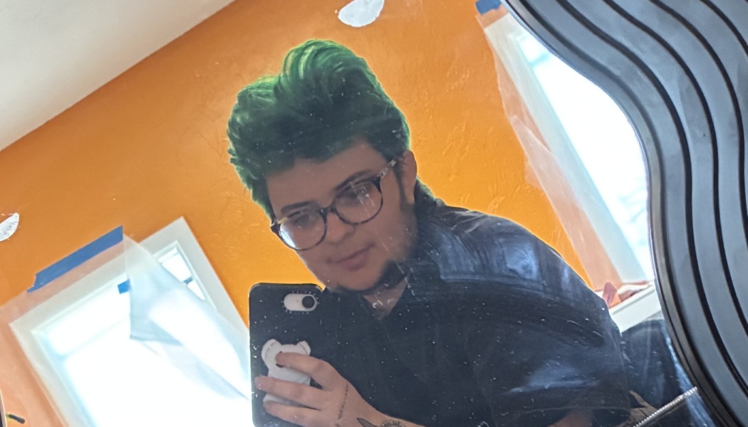 Person with short green hair and glasses taking a mirror selfie against a bright orange wall.
