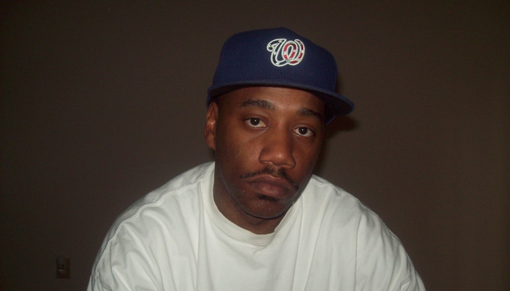 A man wearing a white t-shirt and a navy blue baseball cap looks directly at the camera.