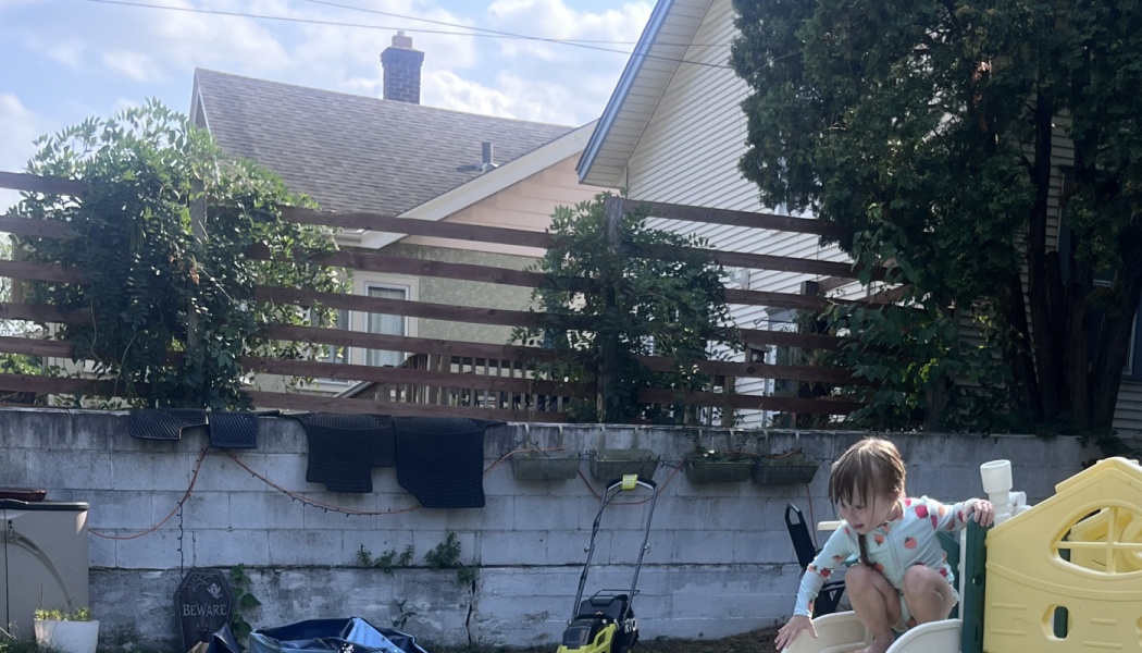 A toddler in patterned pajamas crouches on a plastic play slide in a backyard near a cinderblock wall.
