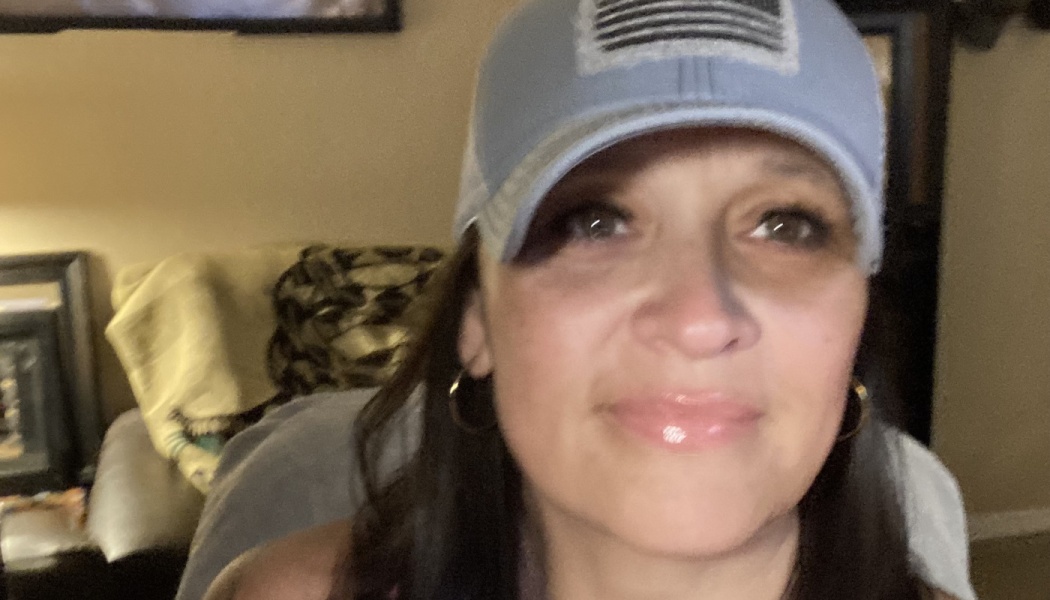 Smiling woman with dark hair wearing hoop earrings and a blue baseball cap with an American flag patch.
