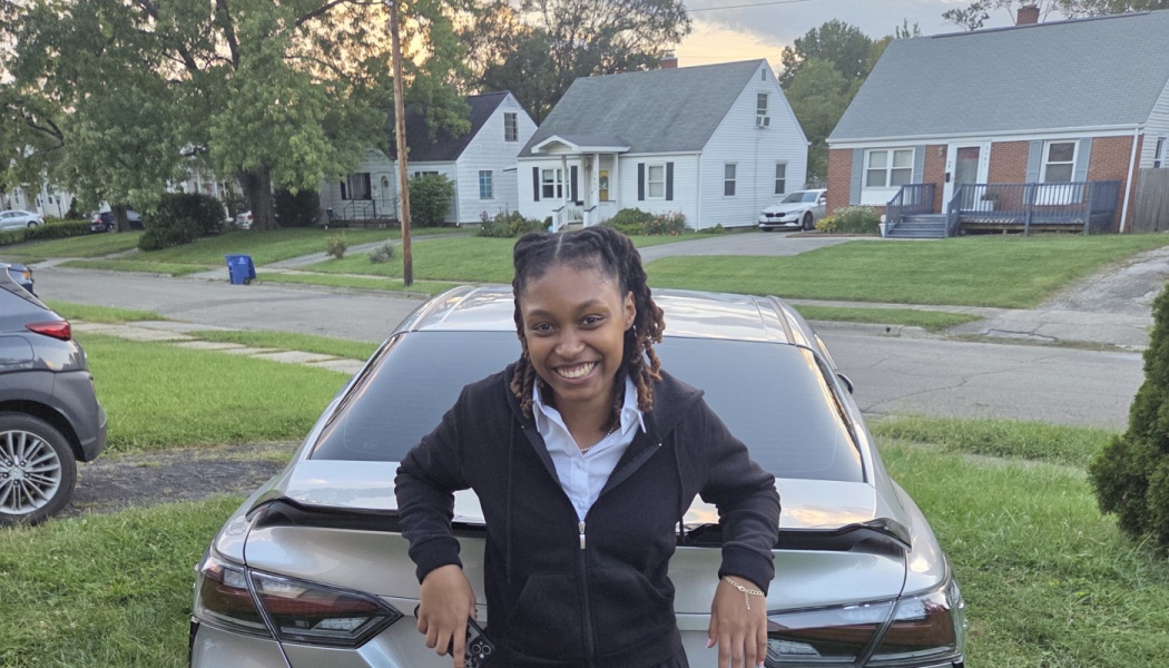 A smiling young woman with locs leans on the trunk of a silver car on a residential street.