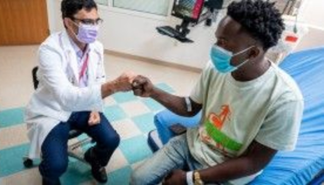 A masked doctor in a white coat and a young male patient share a supportive fist bump in a hospital room.