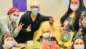A young patient in a mask holds a card, surrounded by supportive healthcare workers making peace signs.