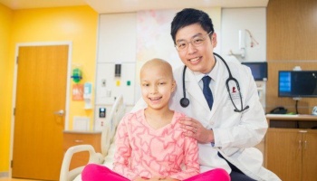 A smiling doctor with his hand on the shoulder of a young bald girl in a hospital room.