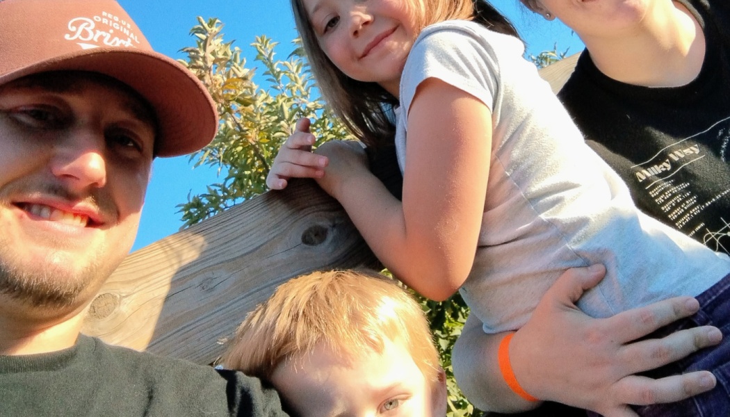 A smiling family of four taking a selfie outdoors near a wooden fence on a sunny day.