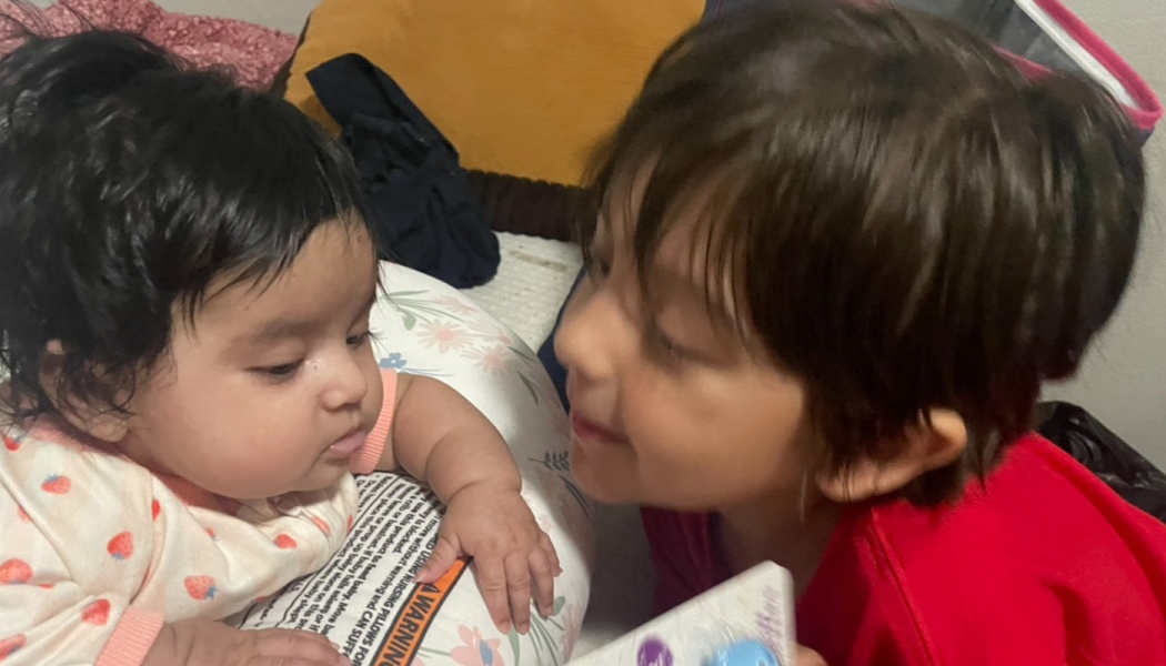 A smiling young child in red leans face-to-face close to a dark-haired baby resting on a support pillow.
