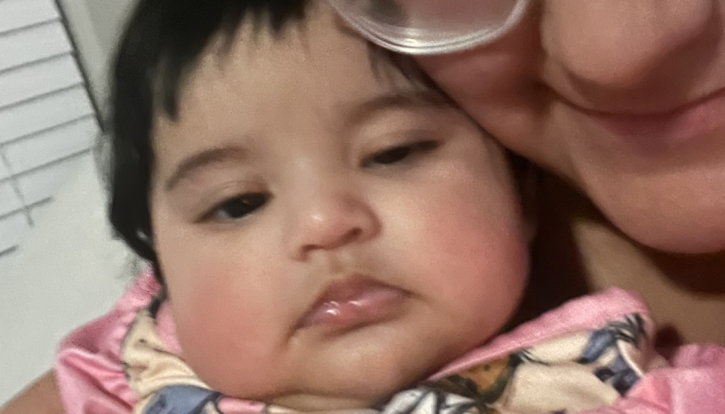 Close-up of a dark-haired baby with chubby, rosy cheeks snuggled against an adult.