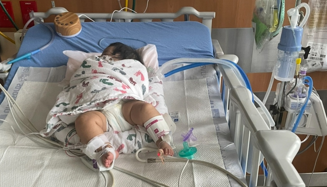 A baby wrapped in a footprint blanket rests in a hospital crib, connected to medical equipment and tubing.