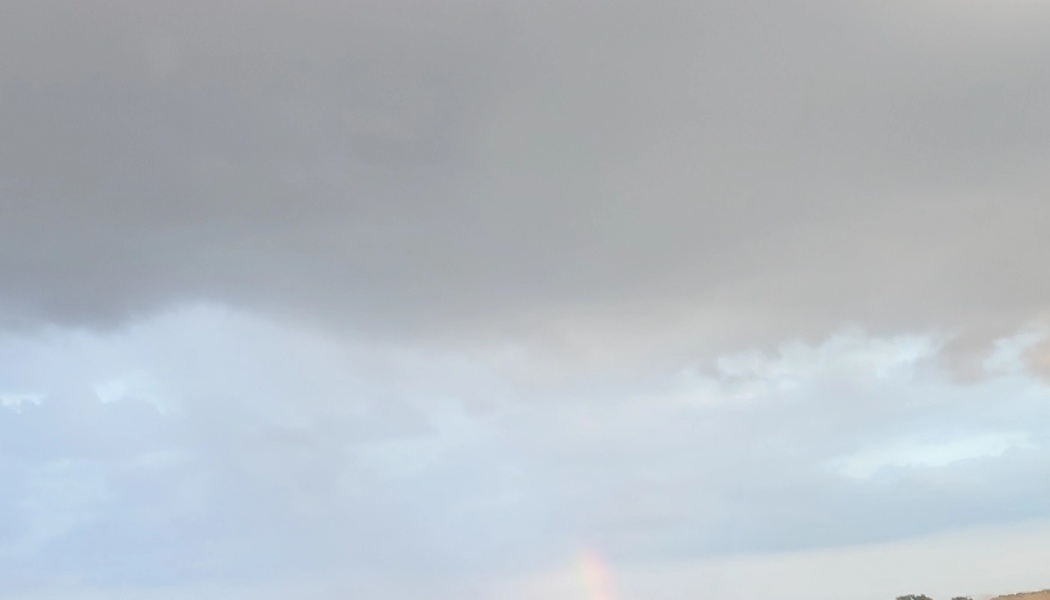 Dark storm clouds fill a pale gray sky, with a faint rainbow appearing near the horizon.