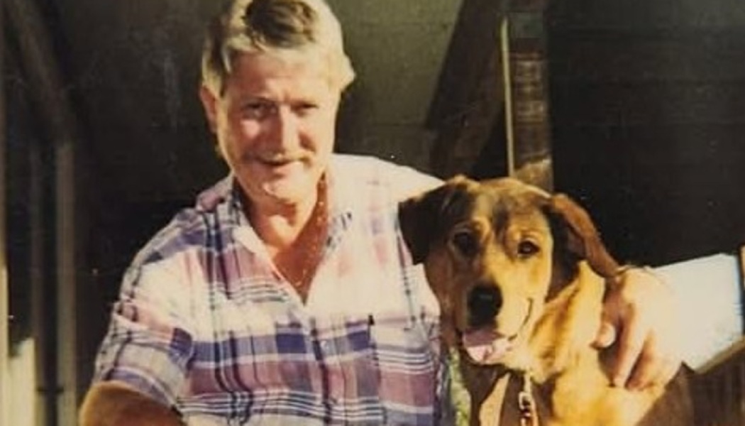 A smiling man in a plaid shirt with his arm wrapped around a happy brown dog.