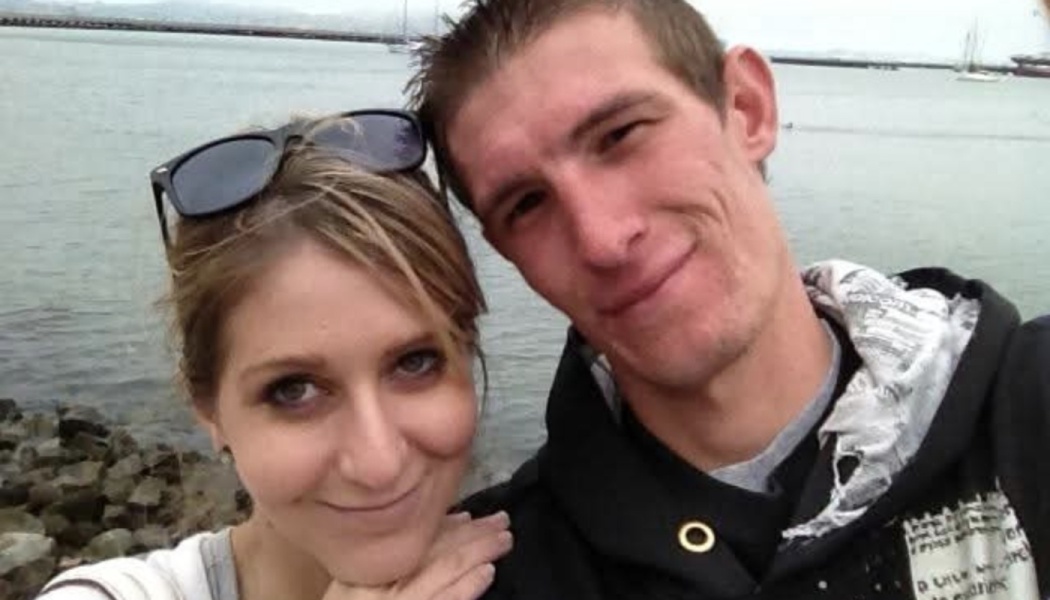 A smiling young couple posing closely for a selfie outdoors near a harbor.