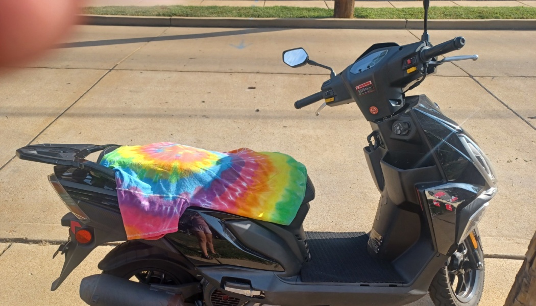 Black motor scooter parked outside with a colorful tie-dye shirt draped over its seat.