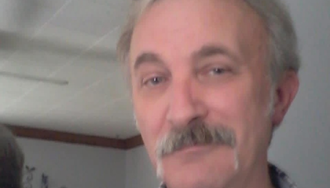 Close-up portrait of a man with graying hair and a thick mustache smiling at the camera.