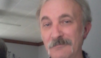 Close-up portrait of a man with graying hair and a thick mustache smiling at the camera.