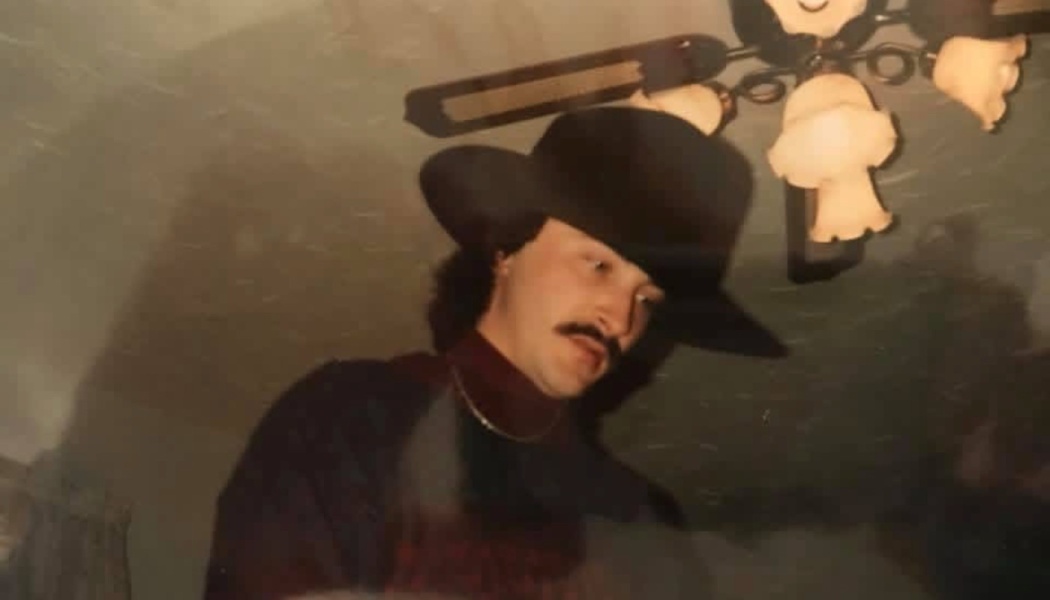 Man with a mustache wearing a black cowboy hat and gold chain, looking downward indoors.