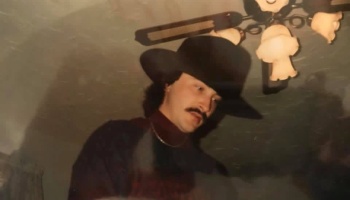Man with a mustache wearing a black cowboy hat and gold chain, looking downward indoors.