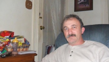Middle-aged man with a mustache wearing a light henley shirt, sitting in an armchair at home.
