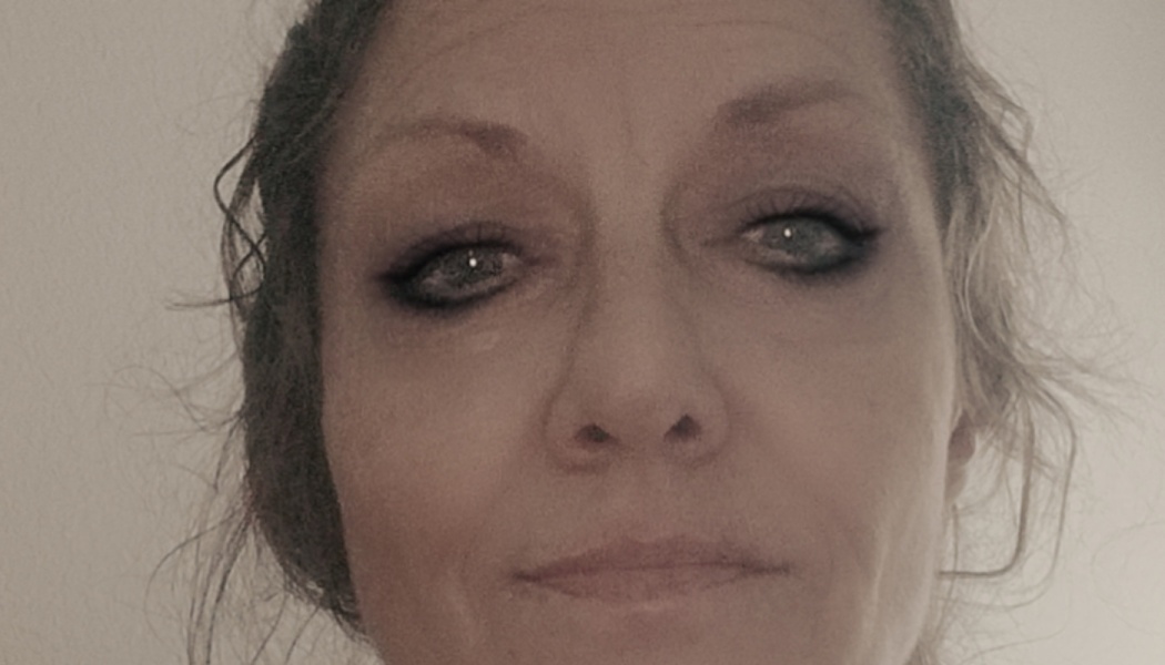 Close-up portrait of a woman with dark eye makeup and light eyes looking directly at the camera.