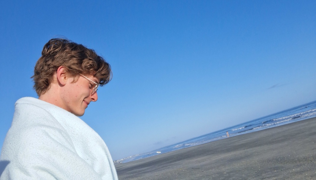 Young man with glasses wrapped in a towel, smiling softly on a sunny beach with the ocean behind him.
