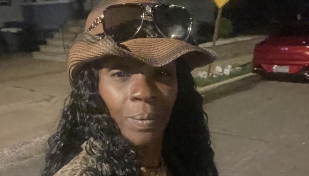 Black woman with long dark curly hair, wearing a brown straw hat with aviator sunglasses resting on the brim.