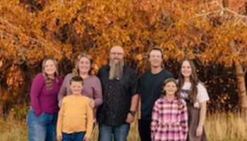 A smiling family of seven standing together outdoors in front of vibrant autumn foliage.
