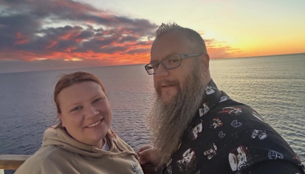 A smiling couple poses on a ship deck with a vibrant ocean sunset in the background.