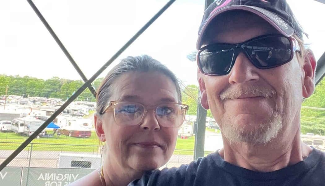 A smiling man wearing sunglasses and a cap taking a selfie with a woman wearing clear-framed glasses outdoors.