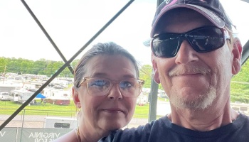 A smiling man wearing sunglasses and a cap taking a selfie with a woman wearing clear-framed glasses outdoors.