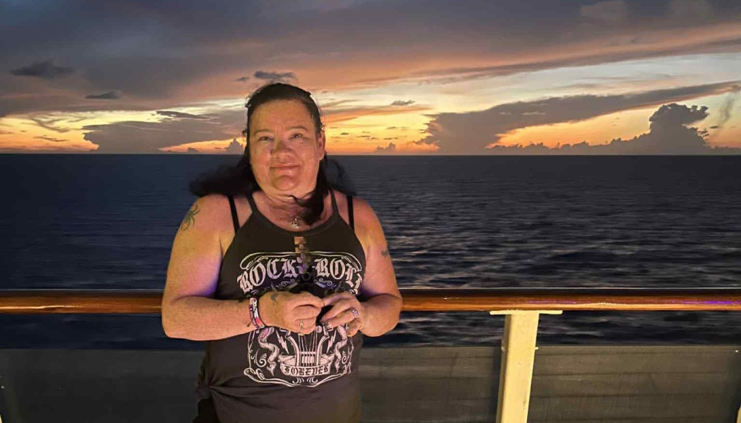 A smiling woman wearing a black graphic tank top stands at a ship's railing in front of a colorful ocean sunset.