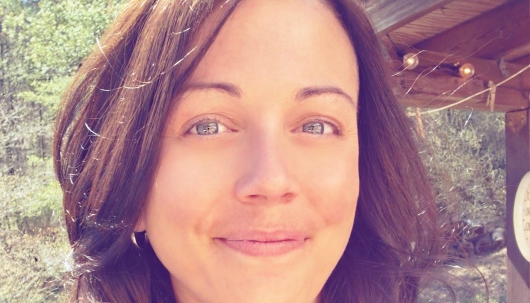 Close-up of a smiling woman with dark brown hair and blue-green eyes outdoors in natural light.