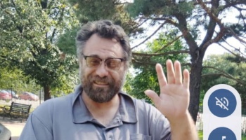 A smiling man with a beard and glasses waving hello outdoors.