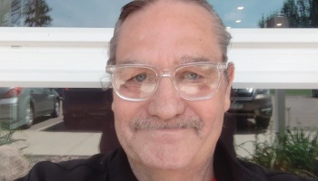Close-up portrait of a smiling man with a mustache and clear-framed glasses.