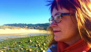 Profile of a woman with glasses and reddish hair looking toward a sunny beach with sand dunes and mossy rocks.