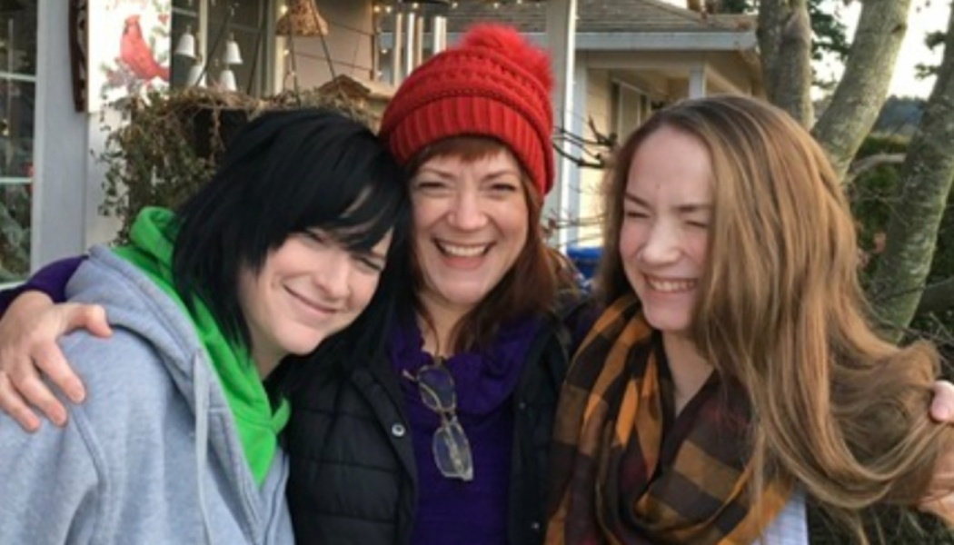 Three happy women smiling and embracing warmly outdoors, one wearing a bright red beanie.
