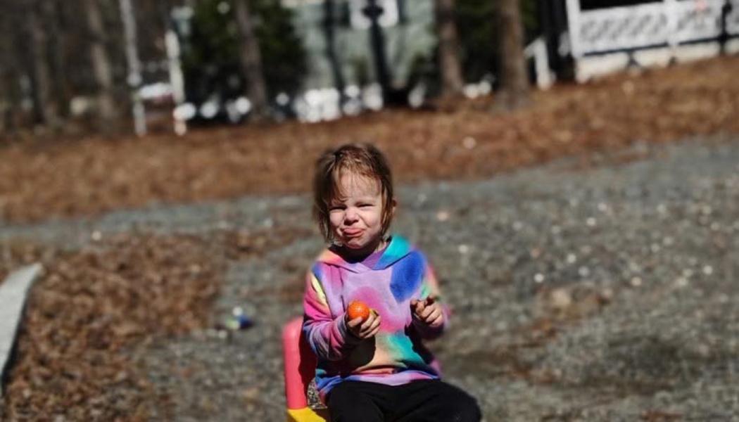 A toddler in a colorful tie-dye hoodie sits outdoors, holding a small orange and making a silly face.