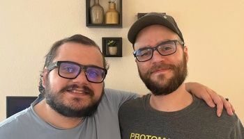 Two smiling men with beards and glasses pose together indoors, one with an arm around the other's shoulder.