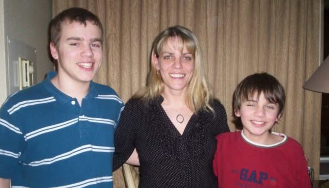 A smiling woman stands with her two sons, posing together happily indoors.