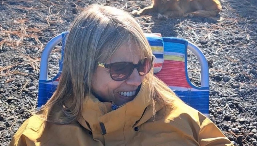A smiling blonde woman wearing sunglasses and a yellow jacket sits in a colorful beach chair outdoors.