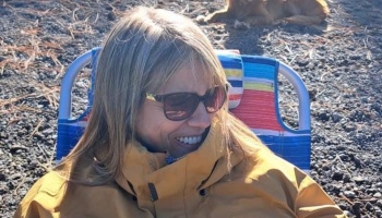 A smiling blonde woman wearing sunglasses and a yellow jacket sits in a colorful beach chair outdoors.