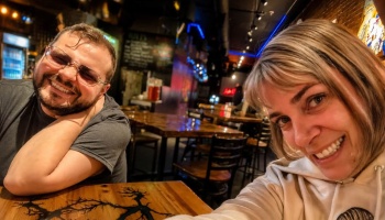 A man with glasses and a woman with blonde bangs smile for a selfie inside a cozy, dimly lit bar.