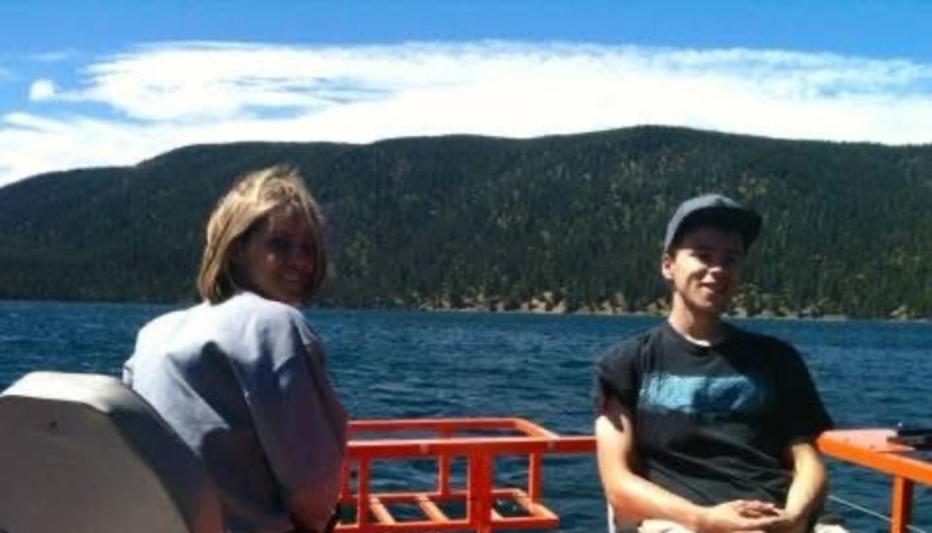 A woman and a young man smiling on a boat with a scenic forested mountain lake in the background.