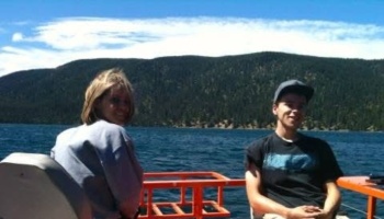 A woman and a young man smiling on a boat with a scenic forested mountain lake in the background.