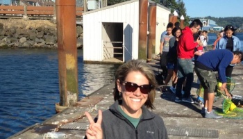Smiling woman in sunglasses gives a peace sign on a sunny dock while people crab in the background.