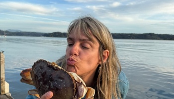 A blonde woman puckering her lips to kiss a large crab she holds by the water.