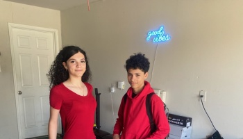 A young woman and teenage boy wearing red stand in front of a blue neon sign that says 'good vibes'.