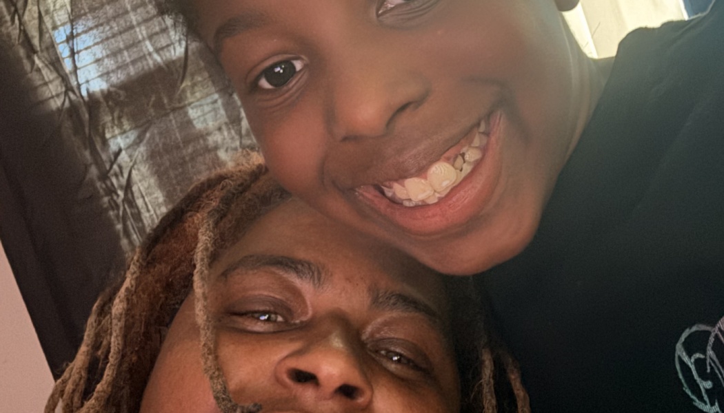 Close-up selfie of a smiling young child posing warmly next to an adult with dreadlocks.